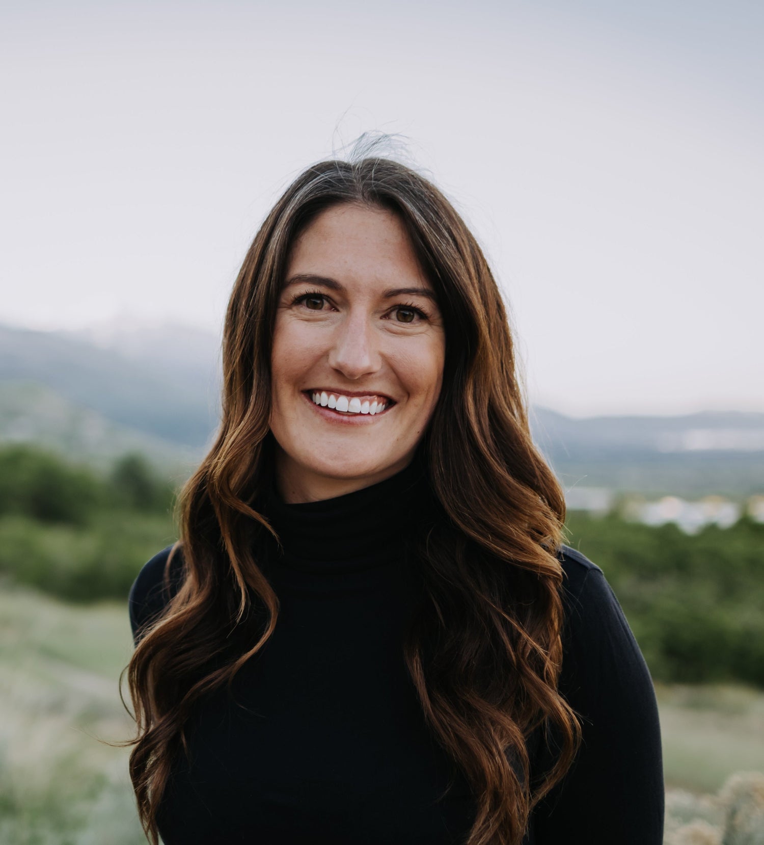 Woman with long brown hair wearing a black turtleneck against a blurred natural background