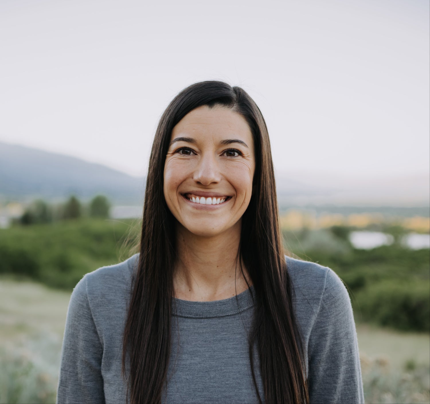 Woman with long dark hair smiling outdoors with mountains in the background