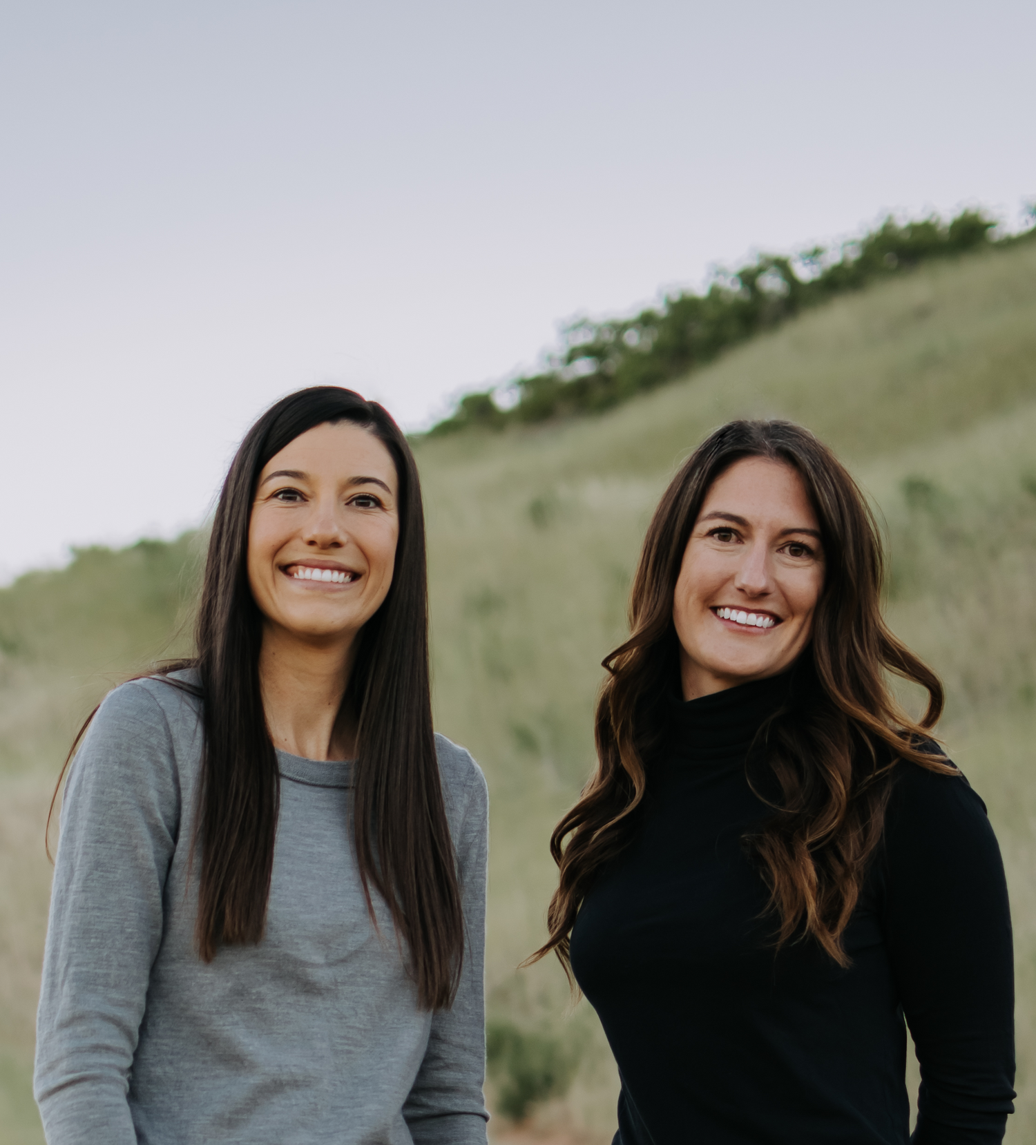 Two women standing outdoors in a grassy hillside landscape, one wearing a gray long-sleeve top and black pants, and the other wearing a black long-sleeve top and dark blue jeans.