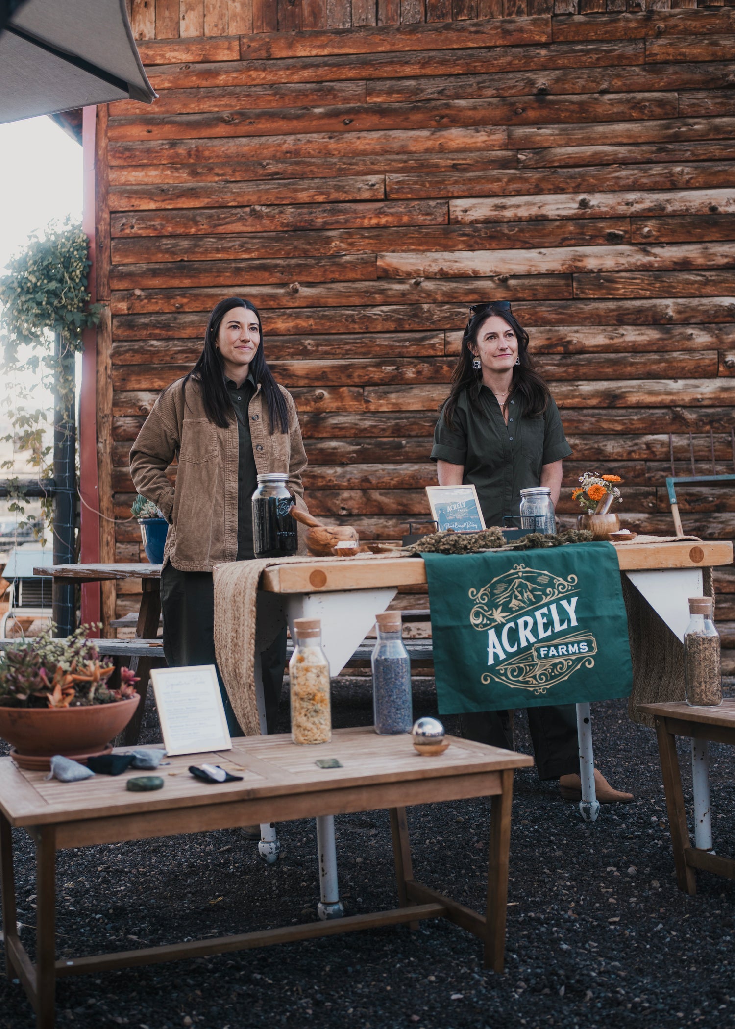 Two women stand behind a rustic wooden table at an outdoor market or farm stand, with a log cabin wall behind them. A green cloth on the table reads 'ACRELY FARMS,' and jars, bottles, and small products are displayed on the tables.