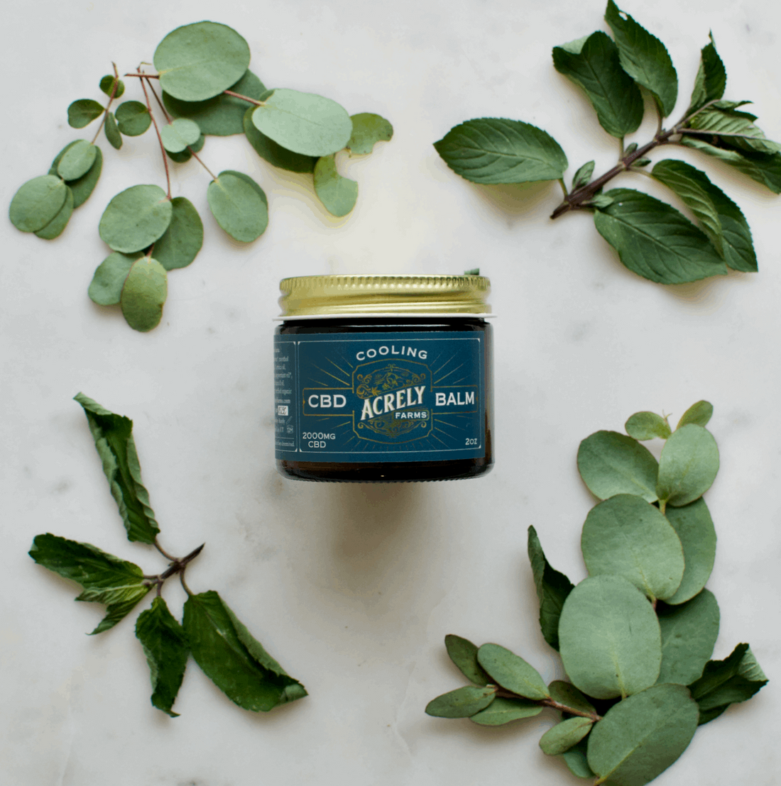 A jar of Acrely Farms Cooling CBD Balm sits on a white marble surface, surrounded by green eucalyptus and mint leaves. The jar has a dark teal label with a gold lid.