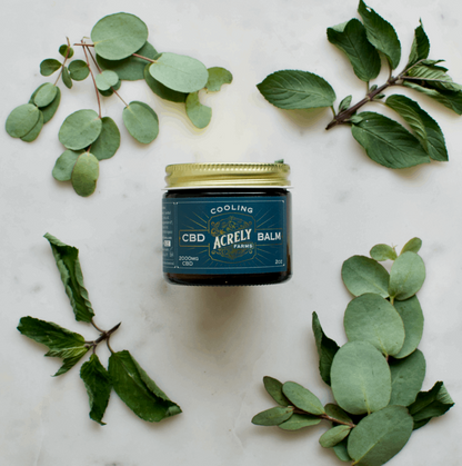 A jar of Acrely Farms Cooling CBD Balm sits on a white marble surface, surrounded by green eucalyptus and mint leaves. The jar has a dark teal label with a gold lid.