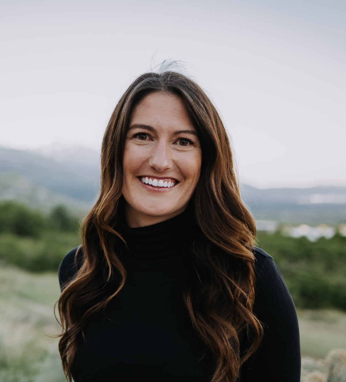 Woman with long brown hair wearing a black turtleneck against a blurred natural background