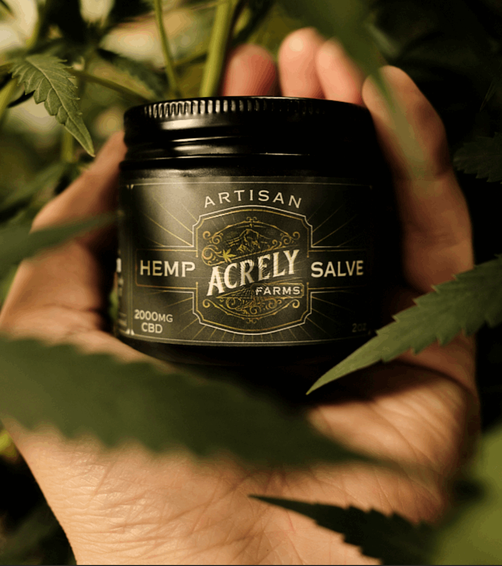 A hand holding a black jar of Acrely Farms Artisan Hemp Salve, surrounded by green cannabis leaves.