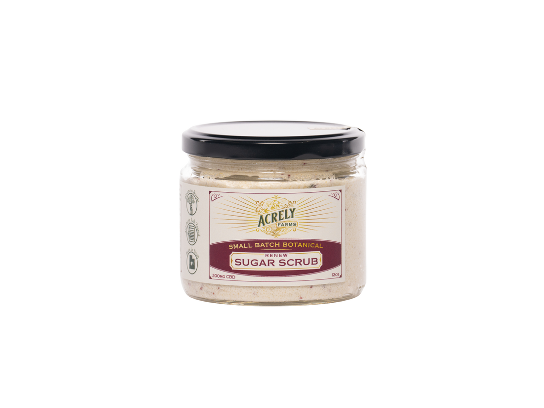 A jar of Acrely Farms Small Batch Sugar Scrub, featuring a beige scrub inside a clear glass jar with a black lid and a cream-colored label.