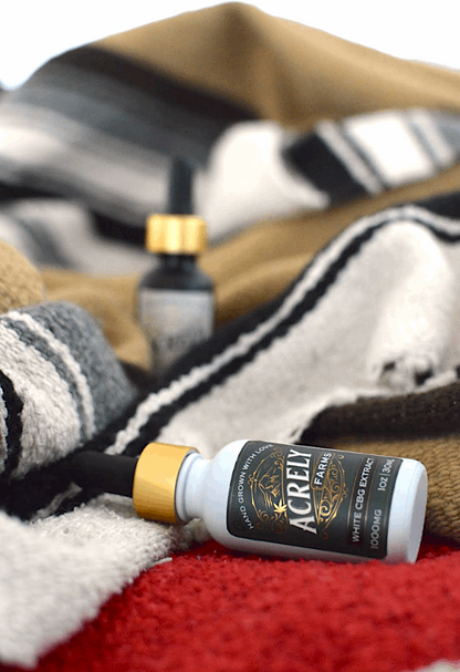 A small white bottle of Acrely Farms White CBG Extract with a gold cap and black dropper lies on a red textured surface, surrounded by folded beige, black, and white striped blankets.