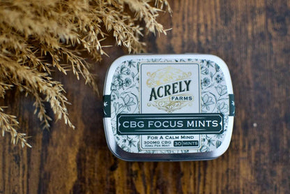 A tin of Acrely Farms CBG Focus Mints placed on a dark wooden surface with dried grass as a decorative prop.
