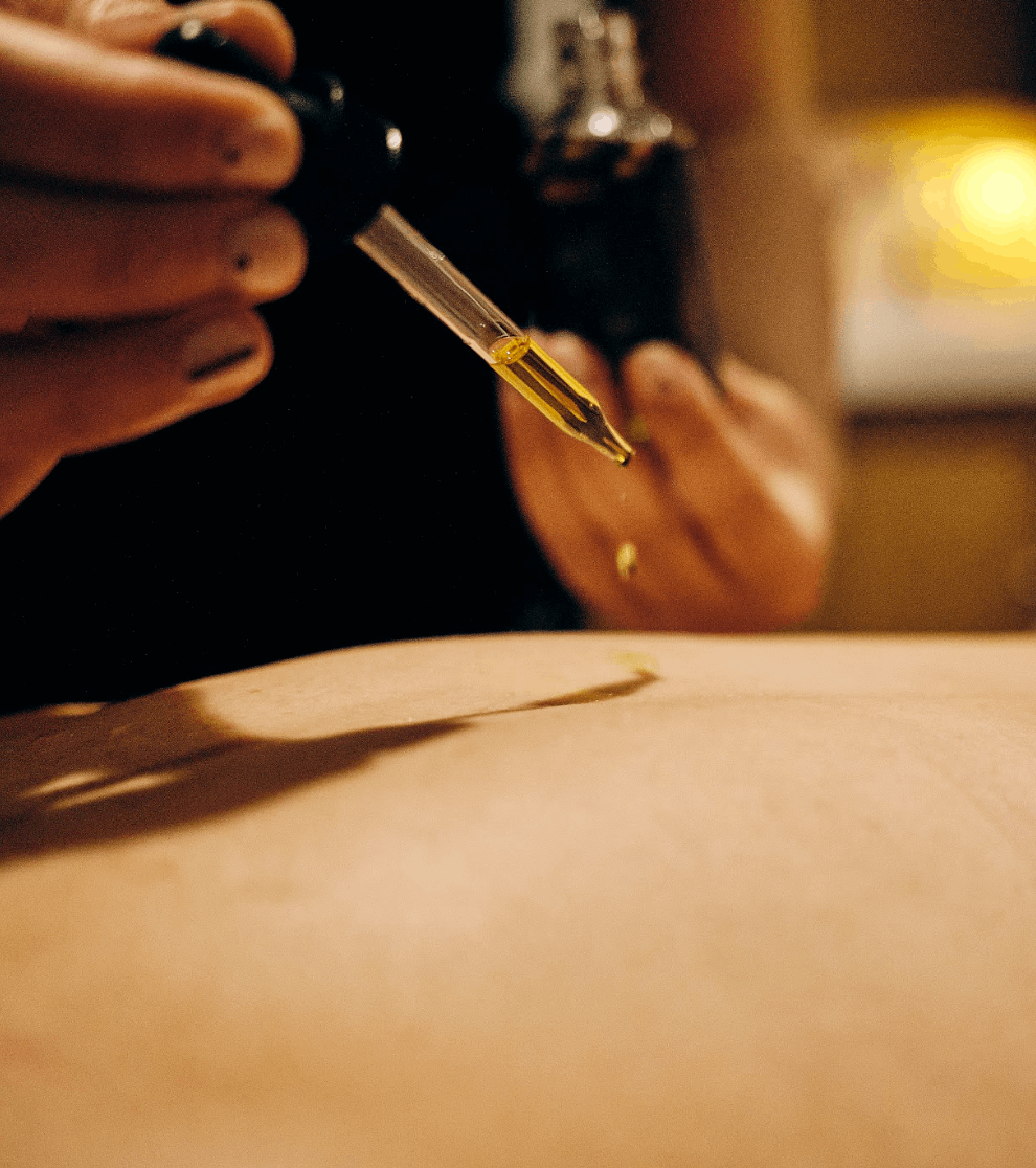 A close-up of a hand holding a glass dropper filled Acrely Farms Gold CBD Body Oil above a skin surface, with a blurred dark bottle in the background.