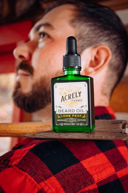 A green glass dropper bottle of Acrely Farms Lone Peak Beard Oil stands on a wooden surface in the foreground, with a bearded man in a red and black plaid shirt blurred in the background.