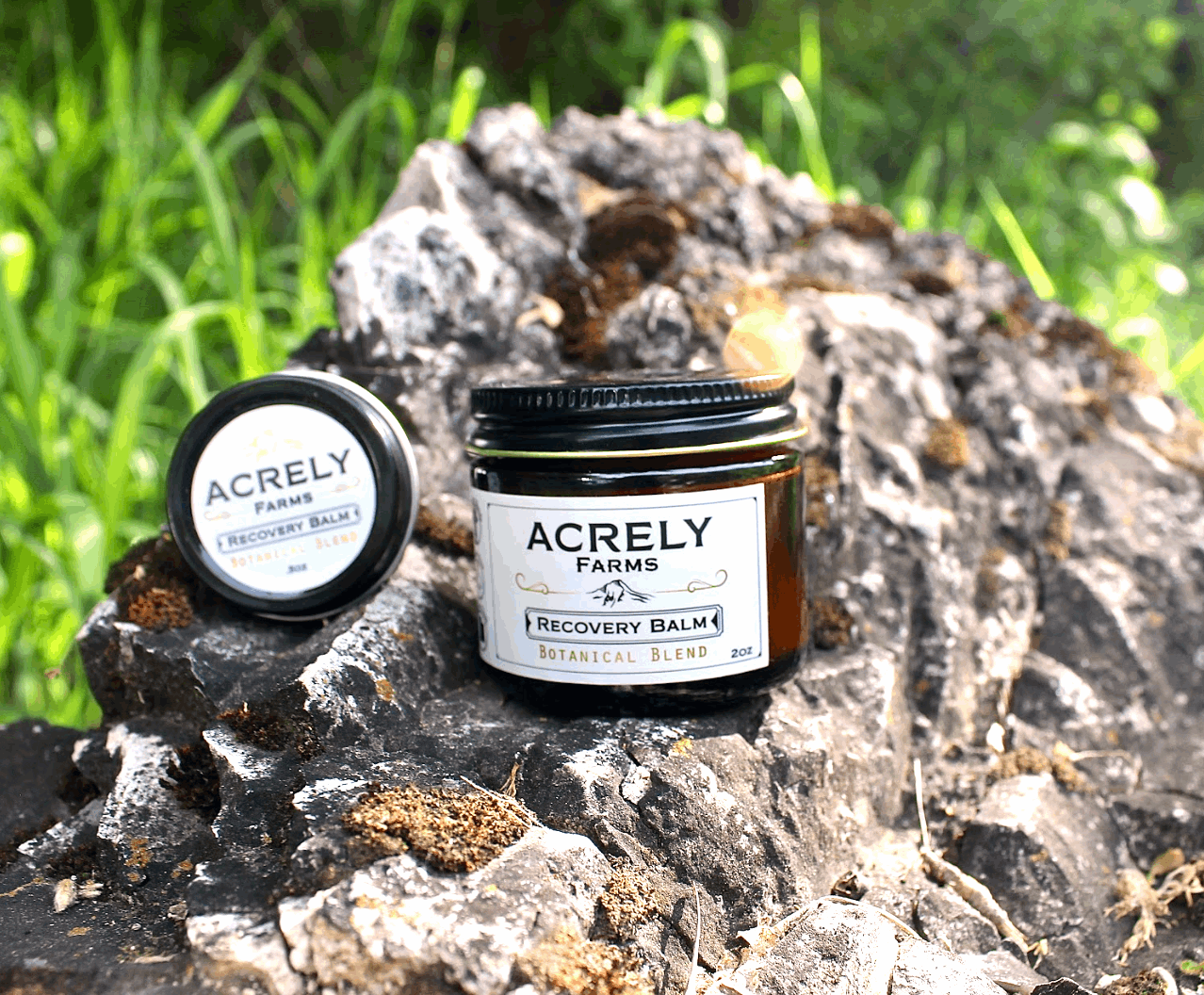 Two jars of Acrely Farms Recovery Balms displayed on a textured rock with a blurred green grassy background.